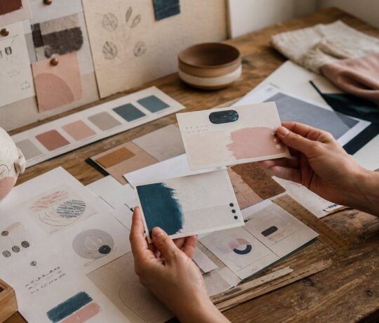 Une créatrice compare deux propositions de cartes sur une table d’atelier couverte de nuanciers, croquis et références visuelles